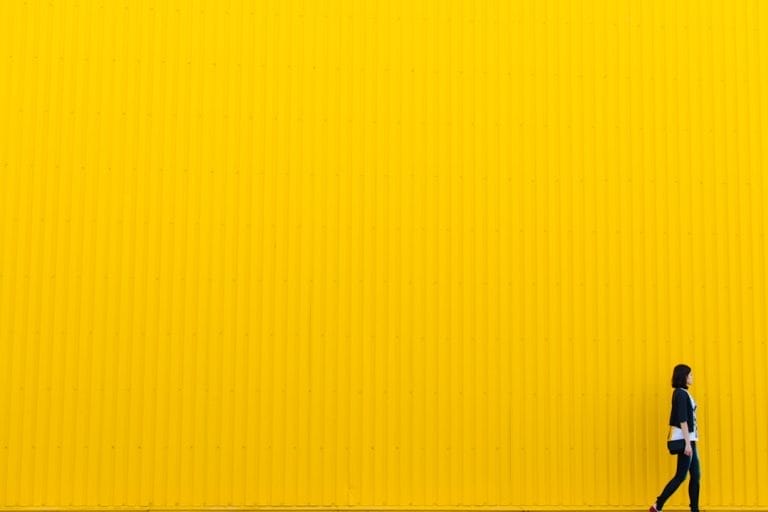 A person walks along the bottom edge of a large yellow wall with vertical lines.