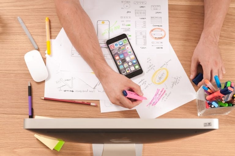 Person highlighting notes on paper with markers at a desk, with a smartphone, mouse, and computer monitor in view.