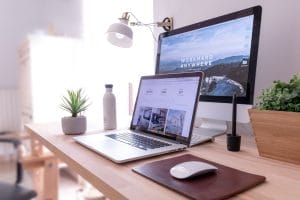 A modern desk setup with a laptop, desktop monitor displaying “WORK HARD ANYWHERE,” a mouse, plant, water bottle, and a desk lamp in a bright workspace.