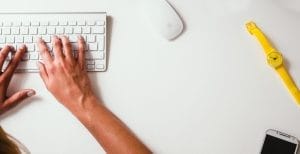 Hands typing on a wireless keyboard next to a computer mouse, a yellow wristwatch, and a smartphone on a white desk.