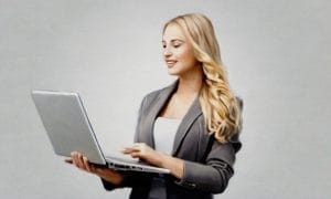 A woman with long blonde hair in a gray blazer is holding and typing on a laptop against a plain light background.