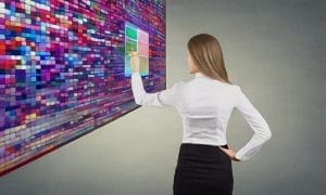 A woman in business attire interacts with a large, colorful digital touchscreen interface displaying various vivid squares and a translucent panel.