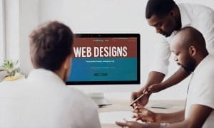 Three men discuss a web design project, with one pointing at a screen displaying a "Web Designs" webpage on a desktop computer.