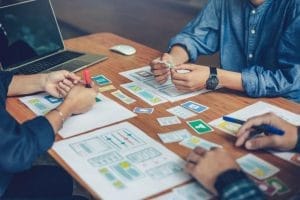 People sitting around a table, working on designing a project with various diagrams and charts. A laptop and stationary tools are also on the table.